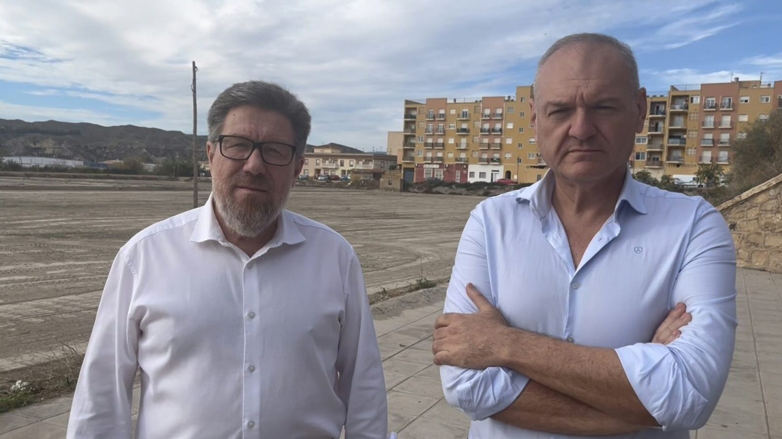 Dos hombres de camisa blanca posan en una explanada de tierra frente a edificios y montañas.