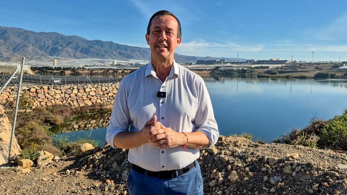 Un hombre de camisa clara posa de pie ante una gran balsa de agua, con montañas al fondo.