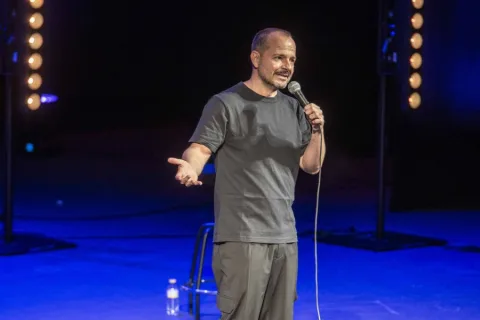 Un hombre calvo con barba, camiseta gris y micrófono en mano, habla en un escenario iluminado en azul.