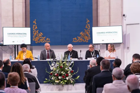Cinco ponentes sentados tras una mesa con un ramo de flores, ante un público y dos pantallas con el título de un congreso internacional.