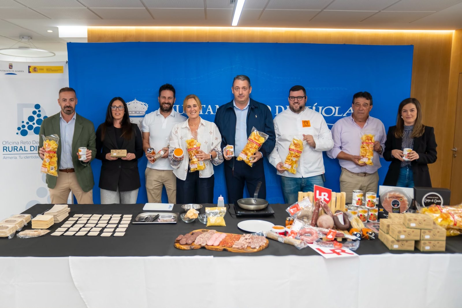 Siete personas de pie tras una mesa con embutidos, quesos, latas y bolsas de snacks, sosteniendo algunos productos ante un fondo azul.