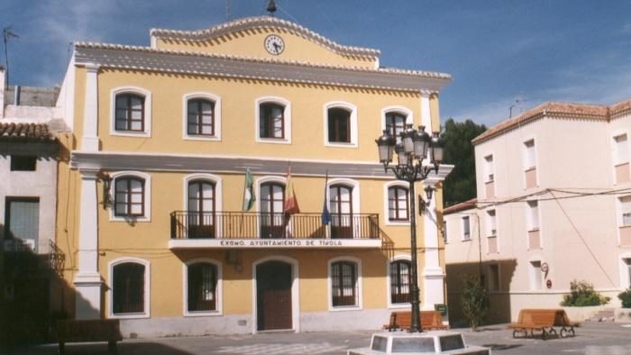 Edificio amarillo de tres plantas con balcón, banderas y reloj en la fachada, junto a una farola y bancos.