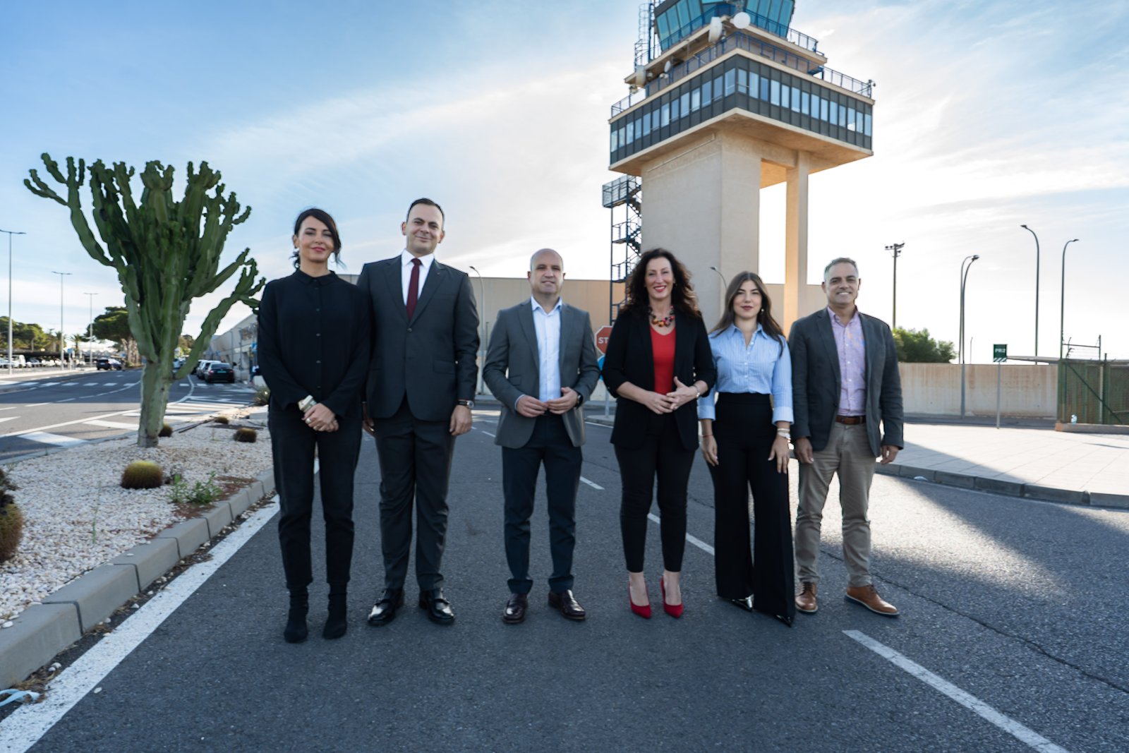 Seis personas trajeadas posan de pie en una calzada junto a una torre de control de aeropuerto.