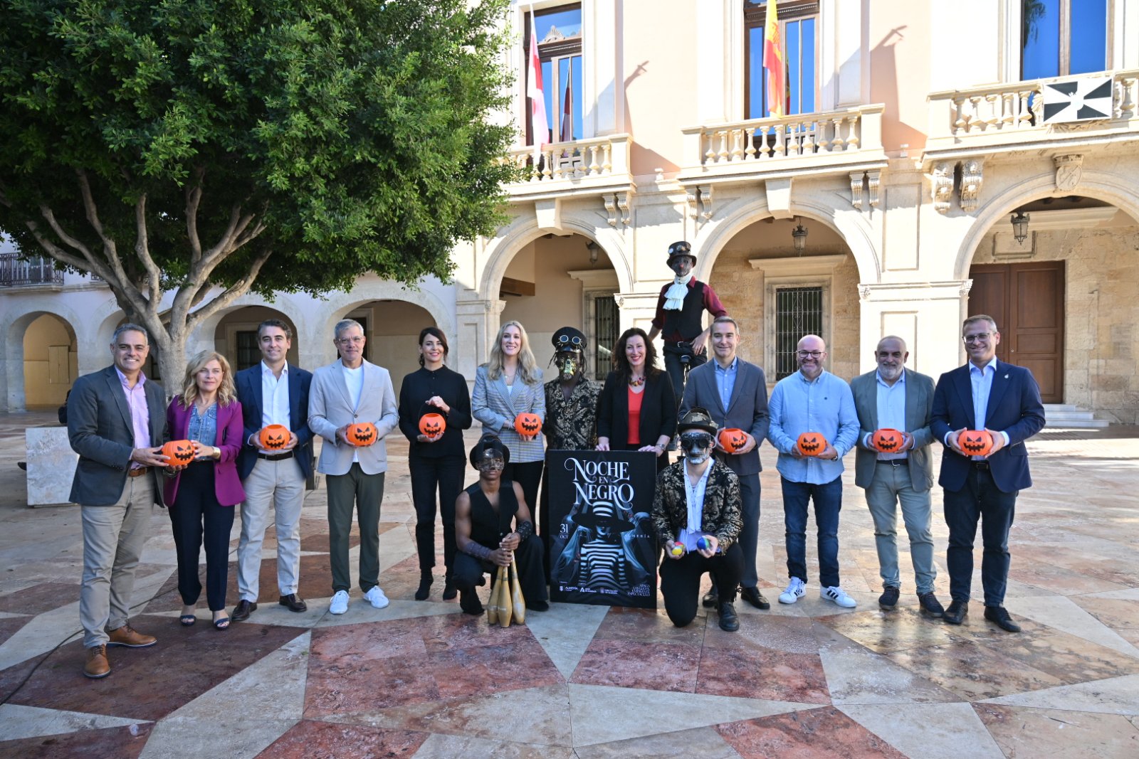 Un grupo de personas posa en un patio con calabazas y un cartel de Noche en Negro, junto a figuras disfrazadas.