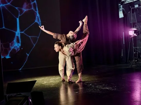 Dos bailarines en escena, uno sostiene a la otra en equilibrio sobre un pie, ante una pantalla con formas azules.