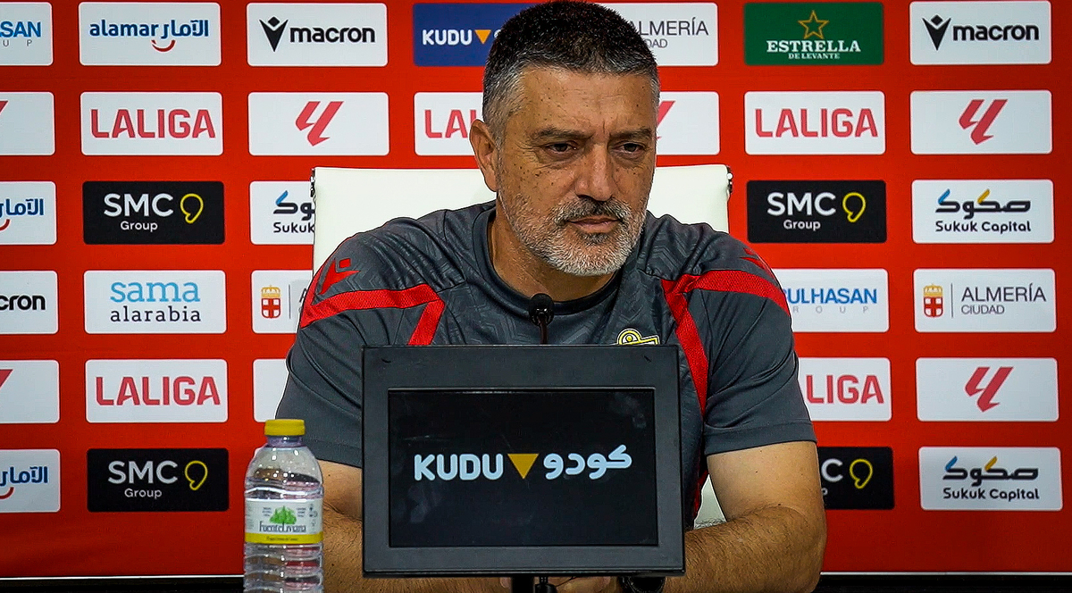 Hombre de barba canosa con camiseta deportiva gris y roja sentado ante un panel rojo de patrocinadores y una pantalla.