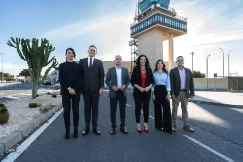Seis personas trajeadas posan de pie en una calzada junto a una torre de control de aeropuerto y un gran cactus.
