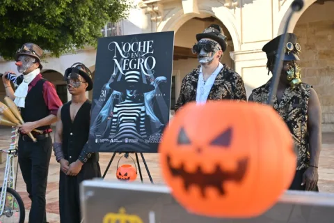Cuatro personas disfrazadas con sombreros y caretas junto a un cartel de Noche en Negro y una calabaza naranja en primer plano.