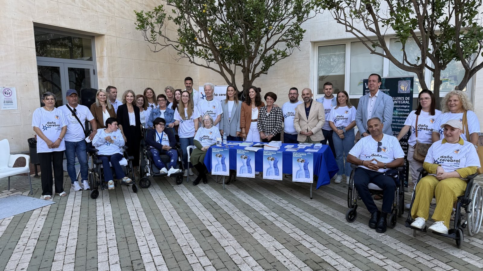 Un grupo de personas, varias en silla de ruedas, posa junto a una mesa con carteles y camisetas de daño cerebral ante un edificio.