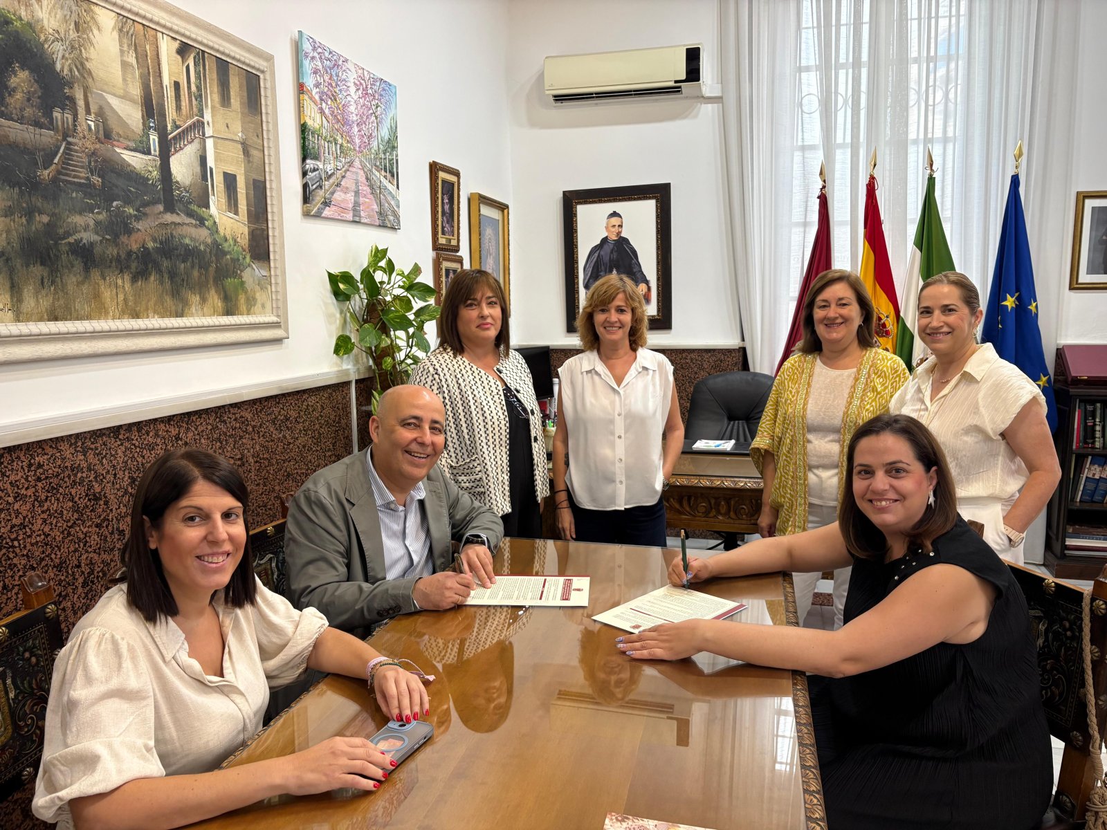 Seis personas posan sonrientes alrededor de una mesa de madera en un despacho con banderas y cuadros, dos de ellas firmando documentos.