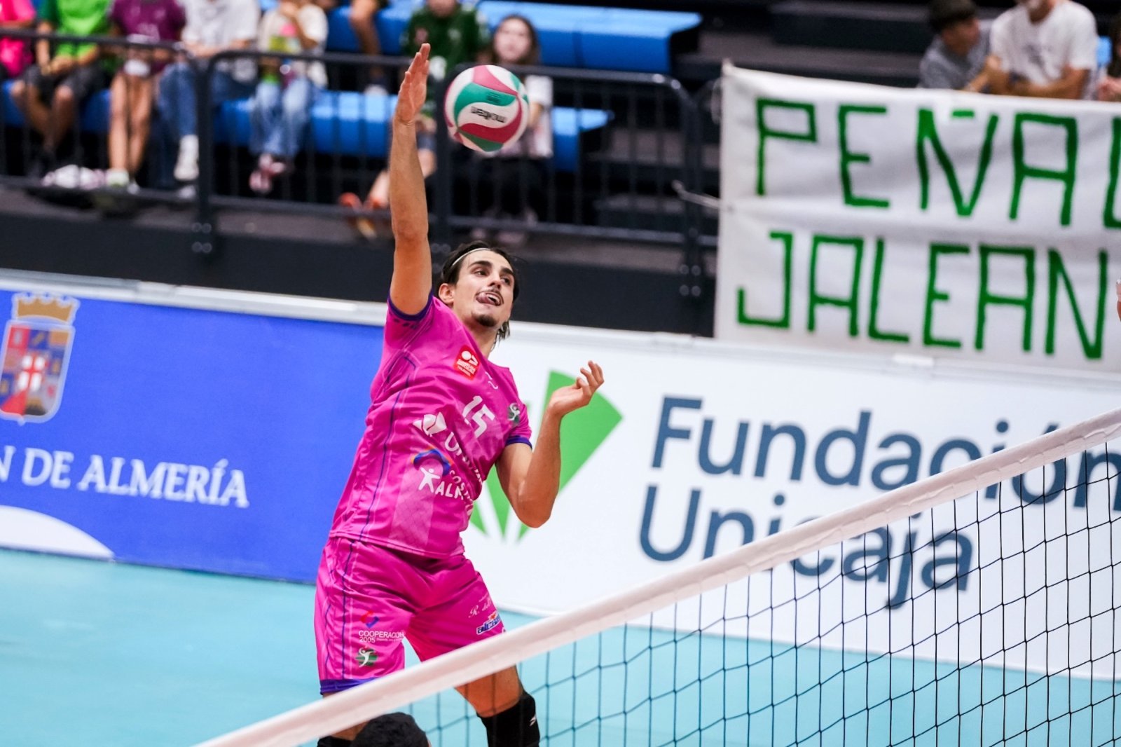Un jugador de voleibol con camiseta rosa salta para golpear el balón junto a la red, ante una grada con público y una pancarta.
