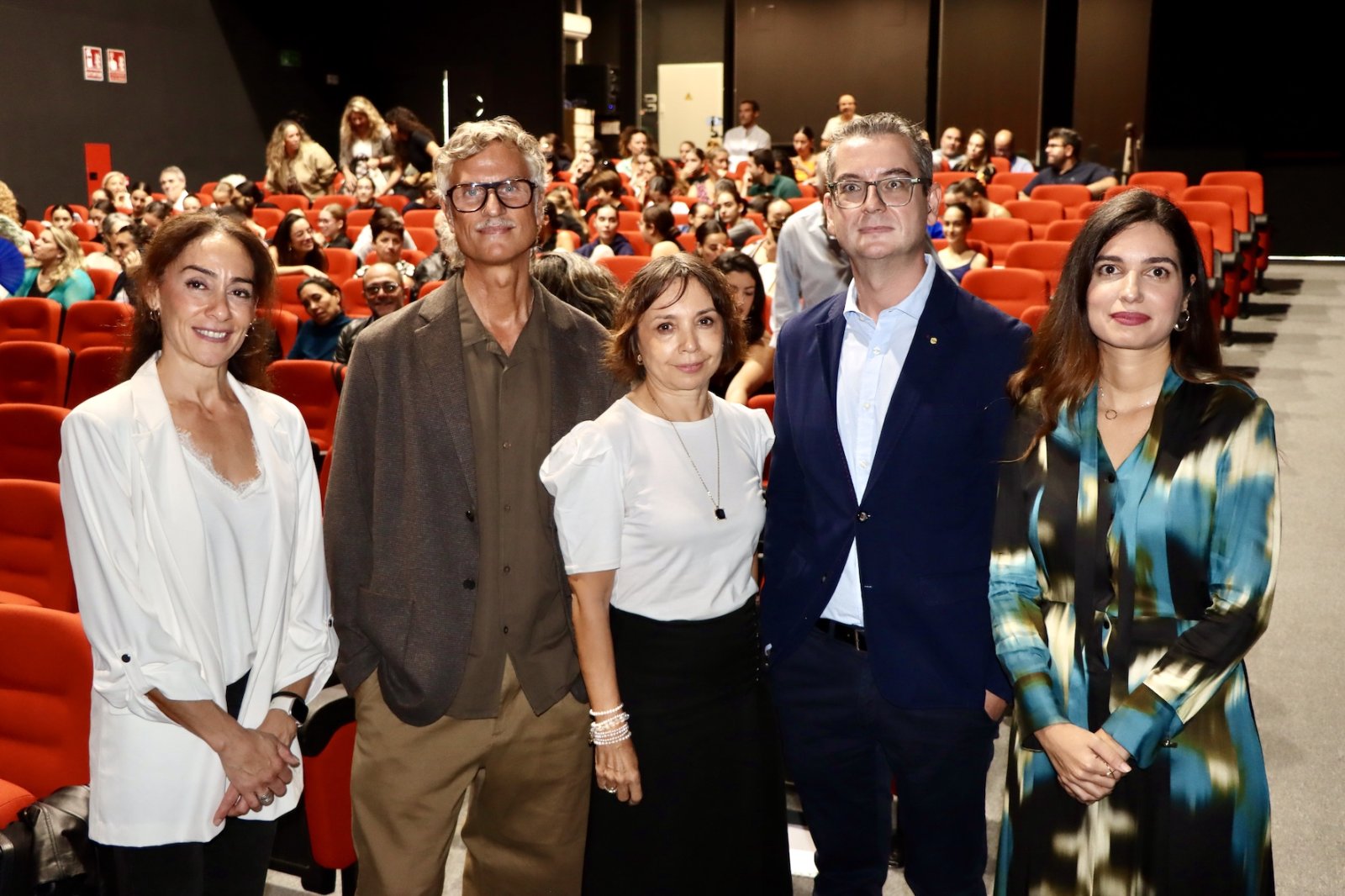 Cinco personas de pie posan en primera fila ante un patio de butacas rojo lleno de público.