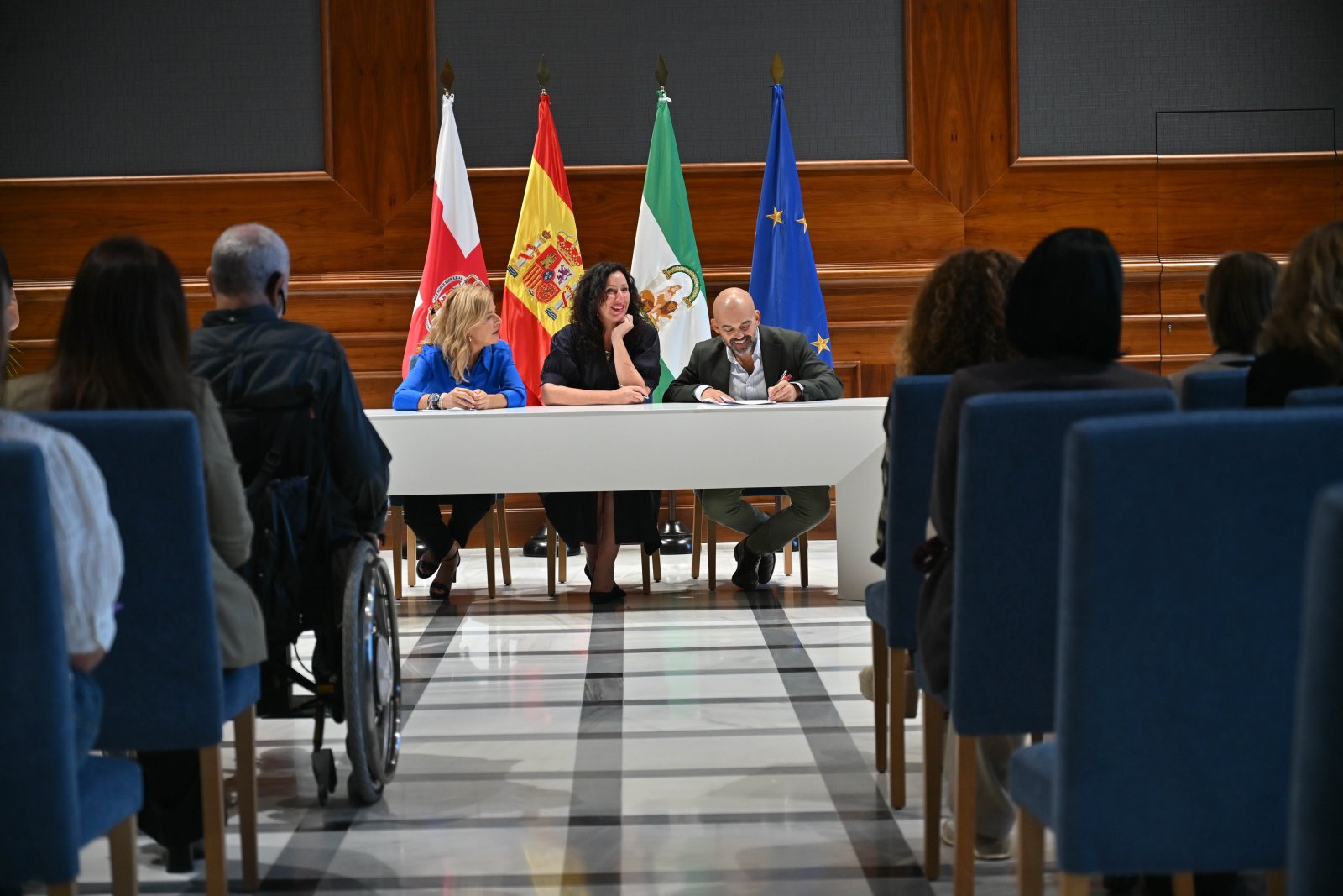 Tres personas sentadas tras una mesa blanca con banderas al fondo, ante un público sentado en sillas azules.