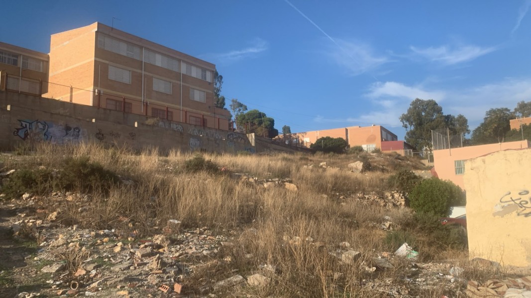 Una ladera con hierba seca y escombros frente a un muro con grafitis y edificios de ladrillo al fondo.