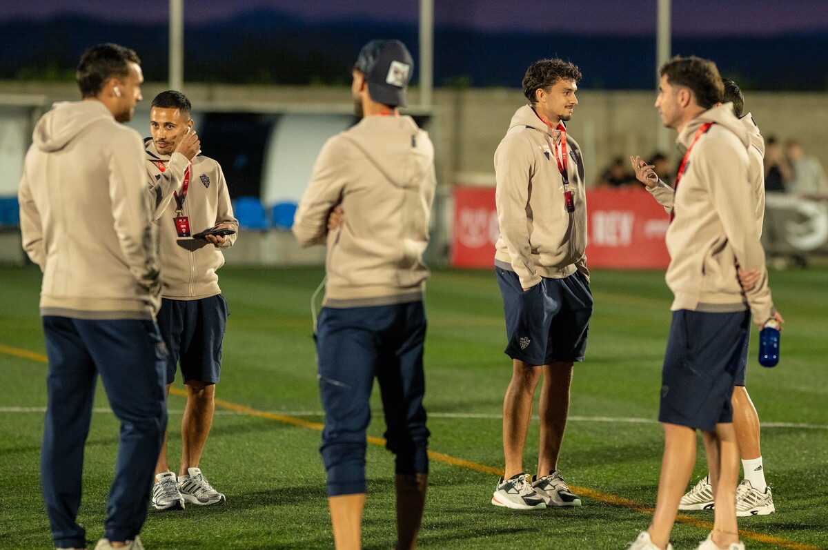 Varios hombres con sudadera beige y pantalón azul conversan de pie sobre un campo de fútbol de noche.