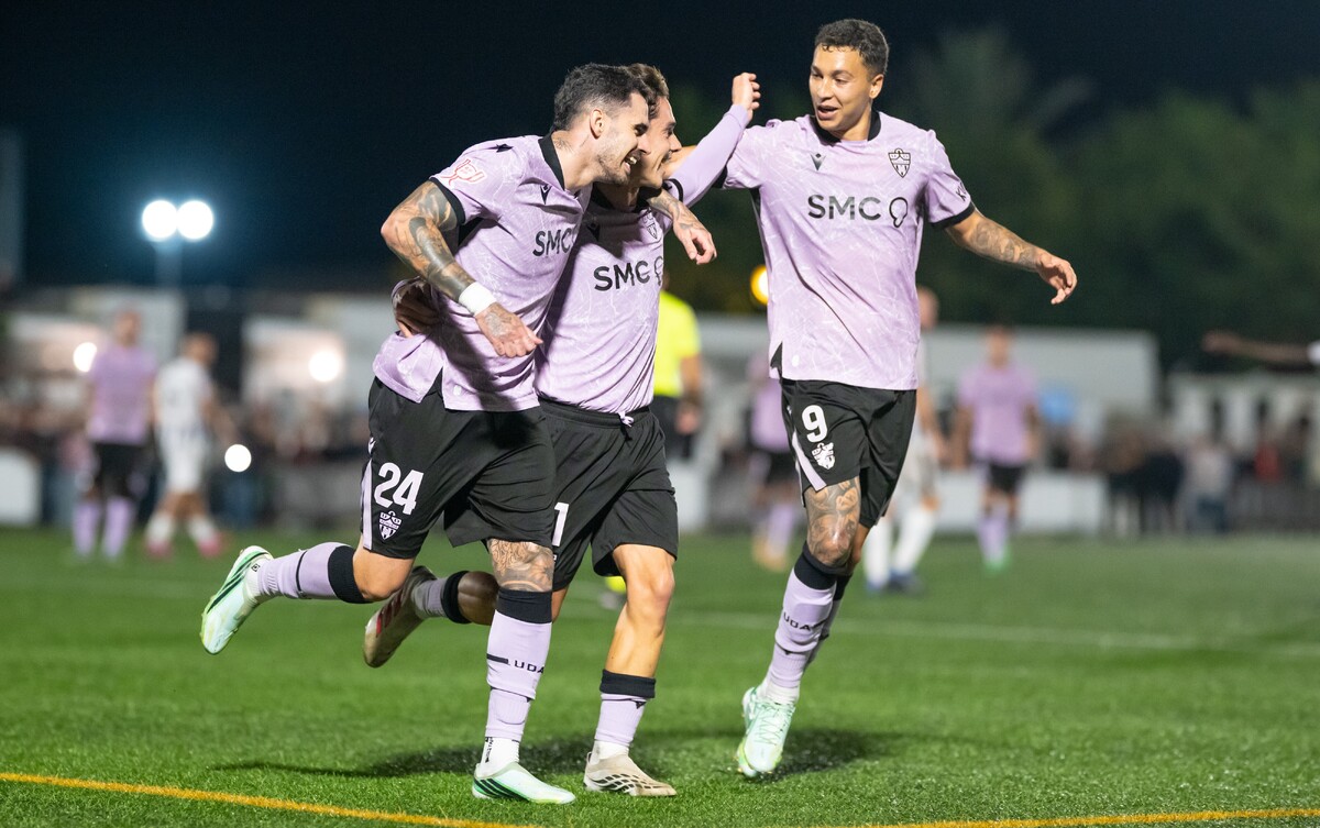 Tres futbolistas con camiseta lila y pantalón negro celebran abrazados sobre el césped de un campo de noche.