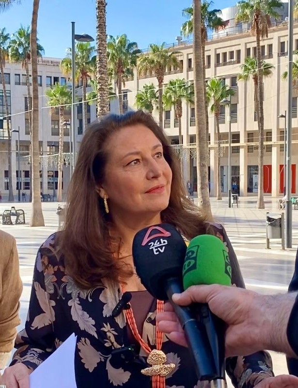 Una mujer de pelo castaño con blusa estampada y collar dorado habla ante varios micrófonos de medios, en una plaza con palmeras y edificios al fondo.