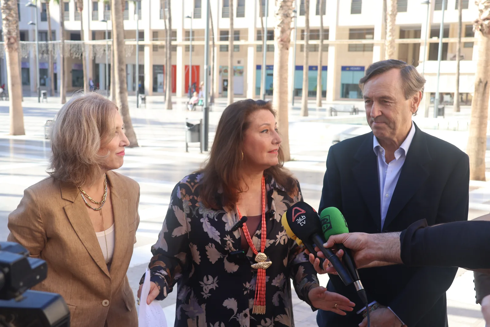 Tres personas conversan al aire libre en una plaza con palmeras, y una de ellas es entrevistada por micrófonos de medios.