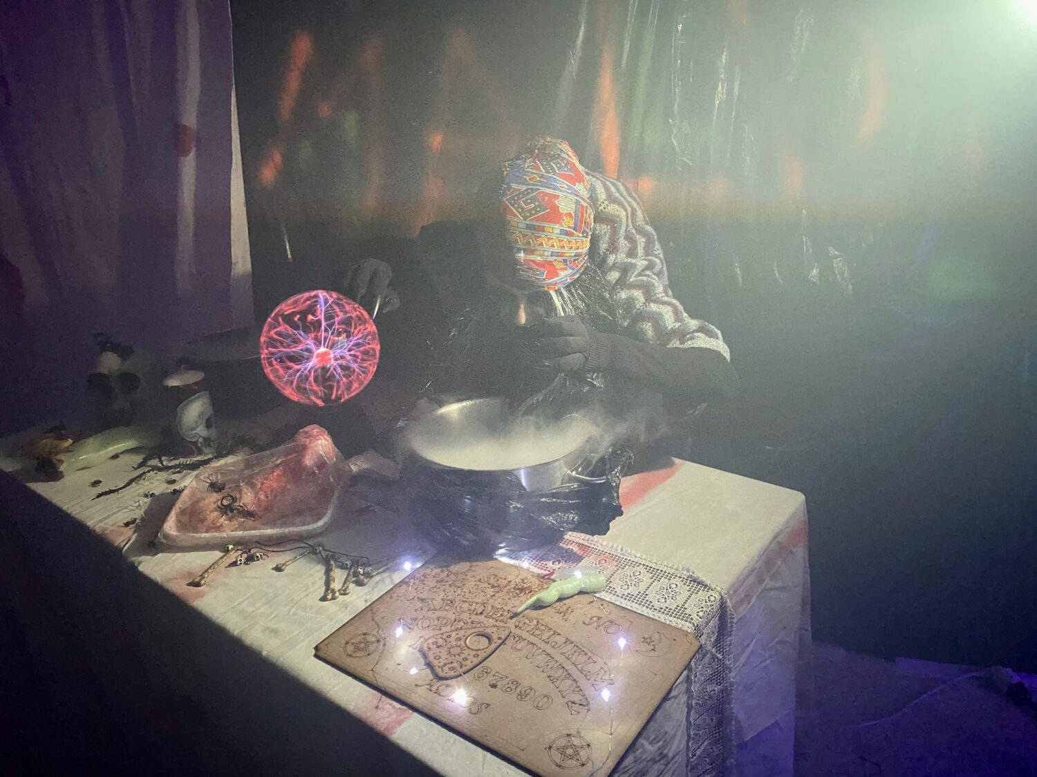 Una persona con el rostro cubierto por un pañuelo estampado manipula una olla humeante junto a una tabla ouija y una esfera luminosa sobre una mesa.