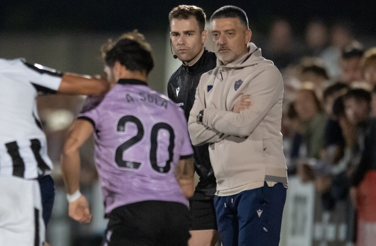 Un entrenador con sudadera beige y brazos cruzados observa junto a un árbitro a un jugador de espaldas con camiseta lila y el número 20.