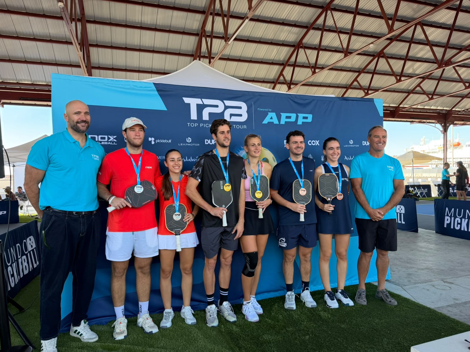 Seis jugadores con palas y medallas posan junto a dos hombres ante un cartel azul de TPS Top Pickleball Tour bajo una cubierta metálica.