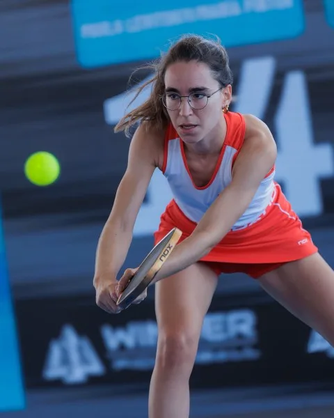 Una jugadora con gafas y ropa naranja y blanca golpea una pelota amarilla con una pala en una pista.