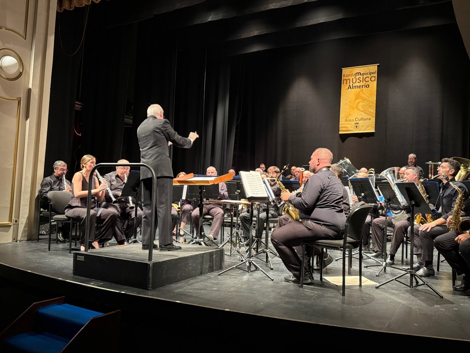 Una banda de música con instrumentos de viento y percusión, dirigida por un hombre de espaldas, sobre un escenario con fondo negro y un cartel colgado.