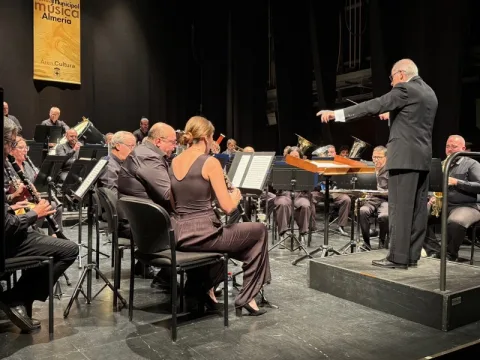 Una banda de música sentada en un escenario con atriles, dirigida por un hombre de traje sobre una tarima.