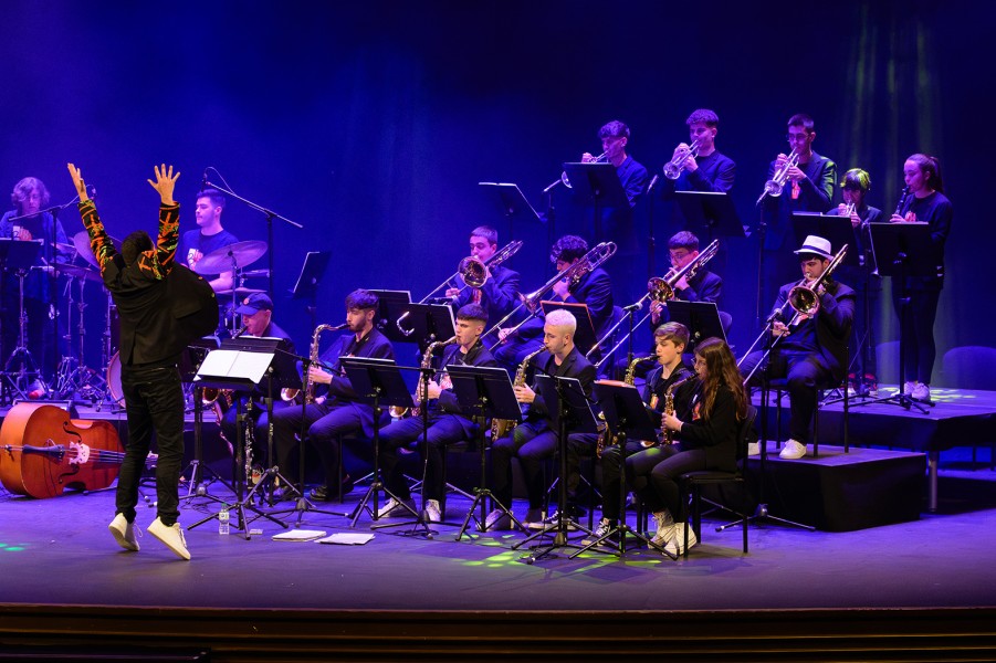 Un director de espaldas con los brazos en alto dirige a una banda de jazz sentada en un escenario con luz azul.