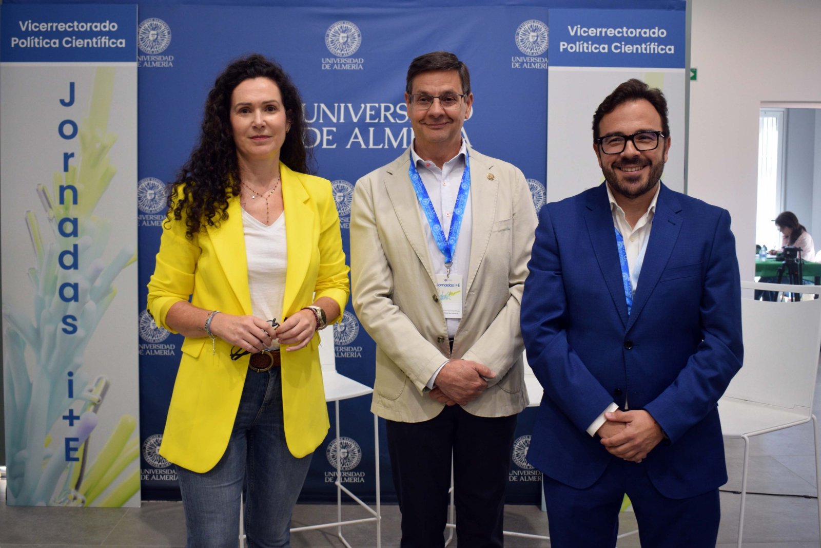 Tres personas de pie posan sonrientes ante un fondo azul con el logotipo de la Universidad de Almería y un cartel de Jornadas i+E.