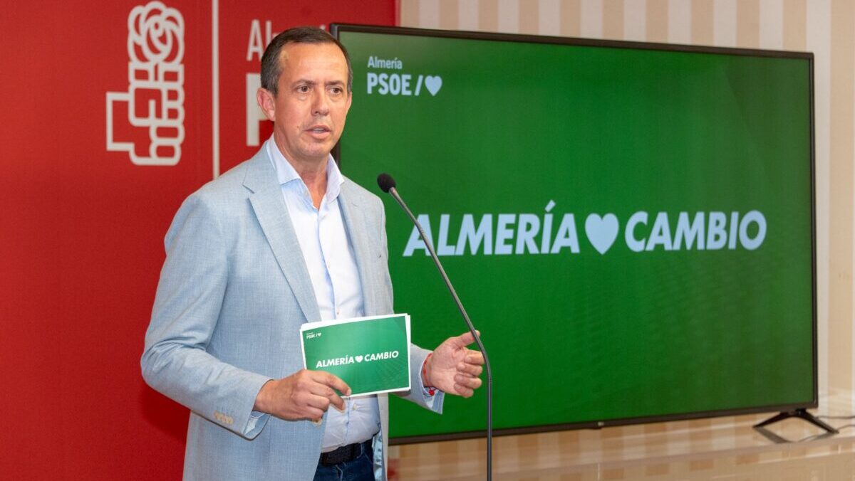 Un hombre con chaqueta azul clara habla ante un micrófono sosteniendo un cartel verde, junto a una pantalla con el lema Almería Cambio.