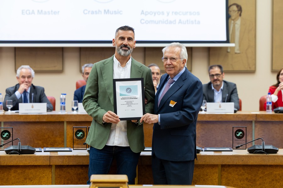 Dos hombres de traje sostienen un diploma enmarcado ante una mesa con micrófonos y una pantalla al fondo.