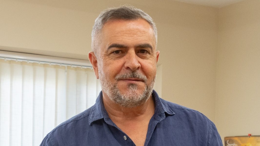 Primer plano de un hombre de mediana edad con barba canosa y camisa azul, en interior.