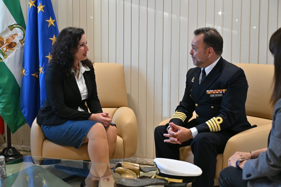 Una mujer y un hombre con uniforme de gala conversan sentados en sillones beige, con banderas de Andalucía y la UE al fondo.