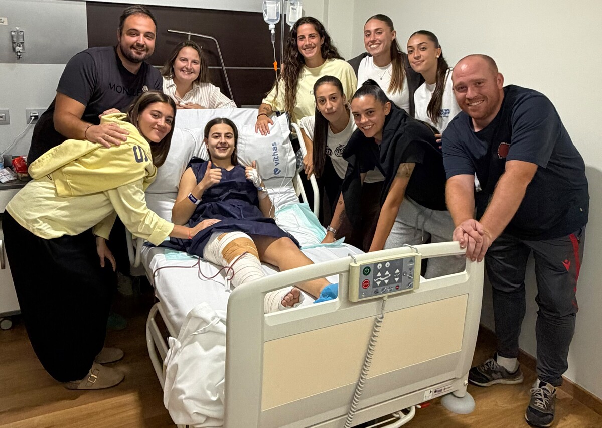 Una mujer joven sentada en una cama de hospital con la pierna vendada, rodeada por ocho personas sonriendo en una habitación clínica.