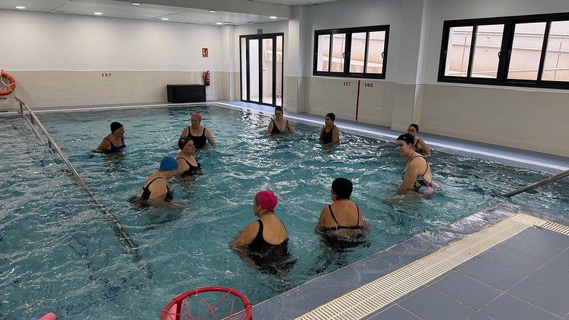 Varias personas con gorro y bañador dentro de una piscina cubierta con ventanales y una canasta de baloncesto.