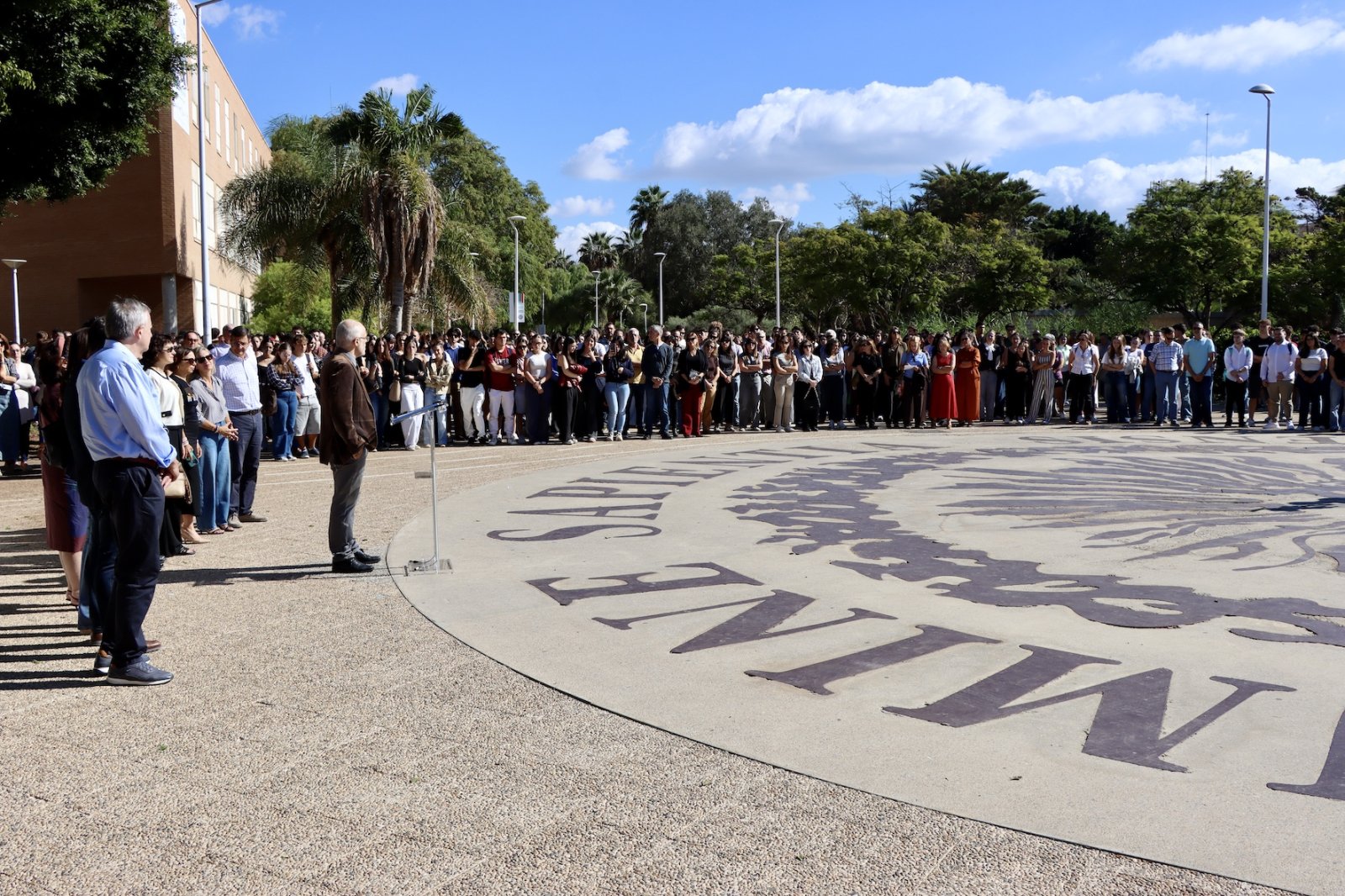 Una multitud en silencio rodea una plaza circular con letras grandes en el suelo, junto a un hombre con un atril.