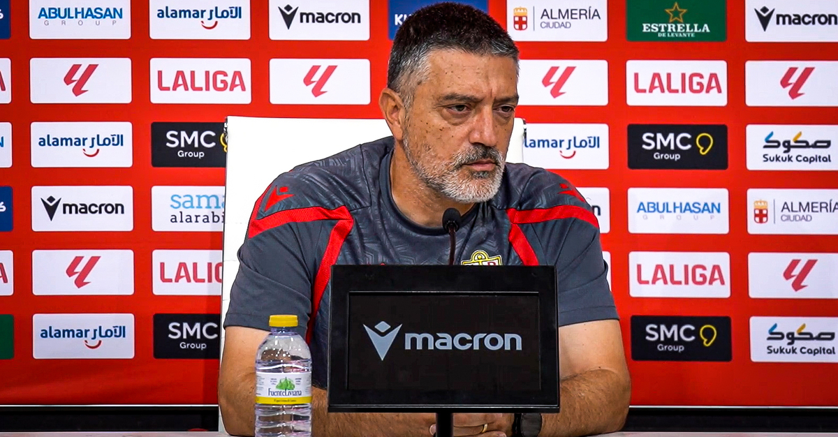 Un hombre de barba canosa con camiseta deportiva habla ante un micrófono en una rueda de prensa, con panel de patrocinadores detrás.