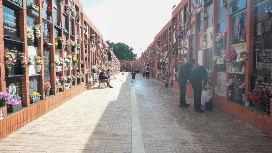 Pasillo de un cementerio entre nichos de ladrillo con flores, con varias personas de pie y una sentada.