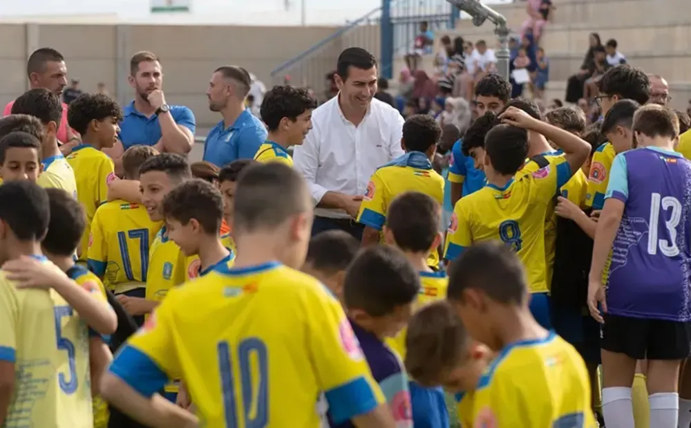 Niños con camisetas amarillas y moradas de fútbol rodean a varios adultos junto a una grada.