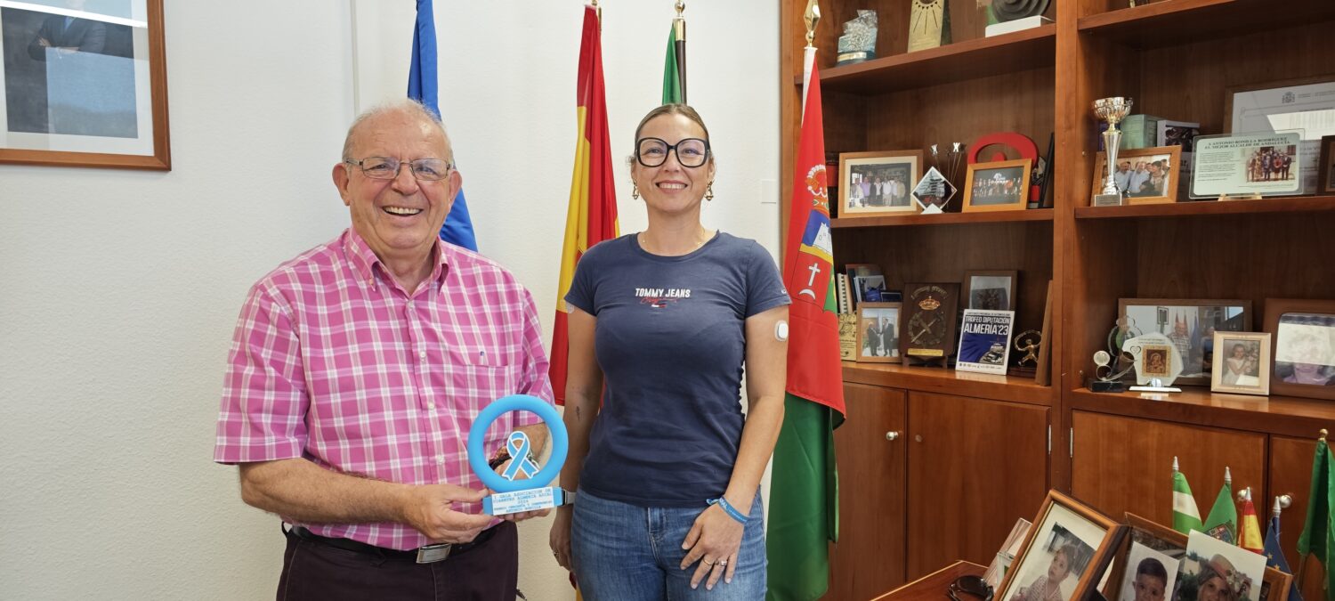 Un hombre mayor con camisa rosa a cuadros y una mujer joven con gafas posan sonrientes junto a banderas y una estantería con trofeos.