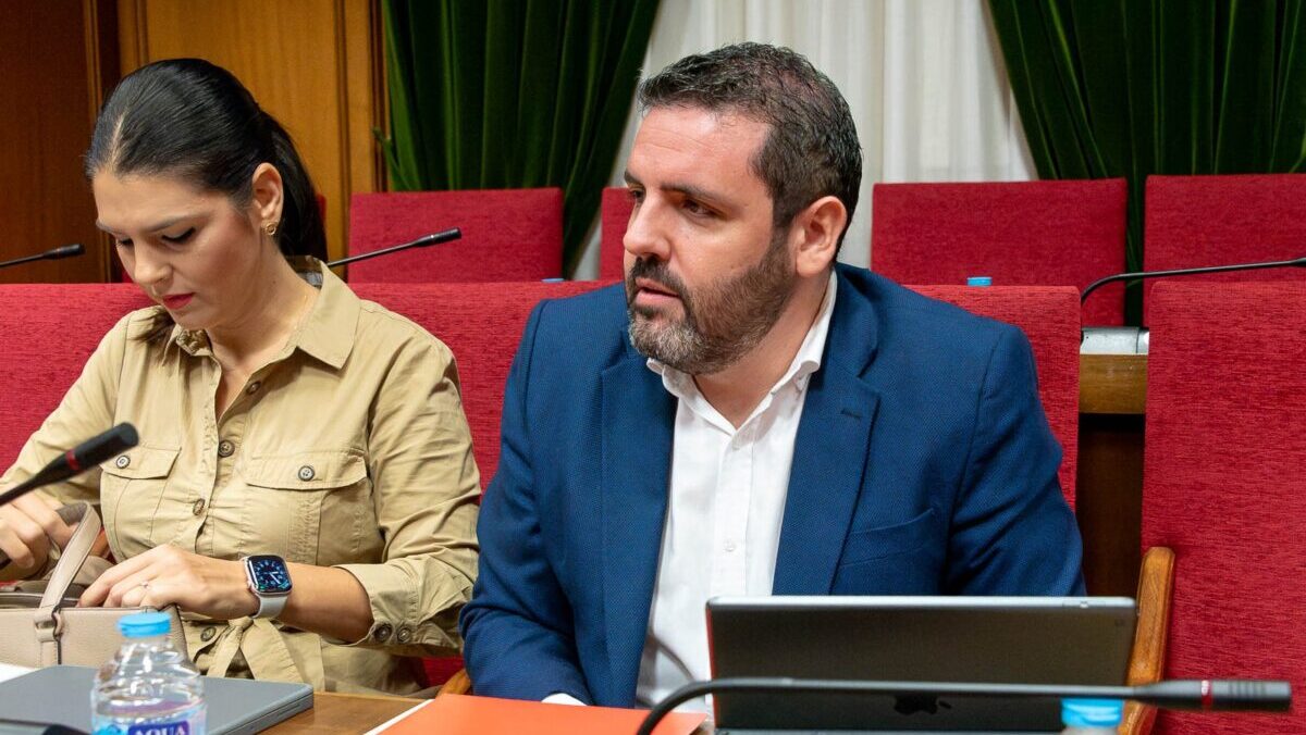 Un hombre con barba y traje azul y una mujer de camisa beige están sentados ante micrófonos en una sala con butacas rojas.