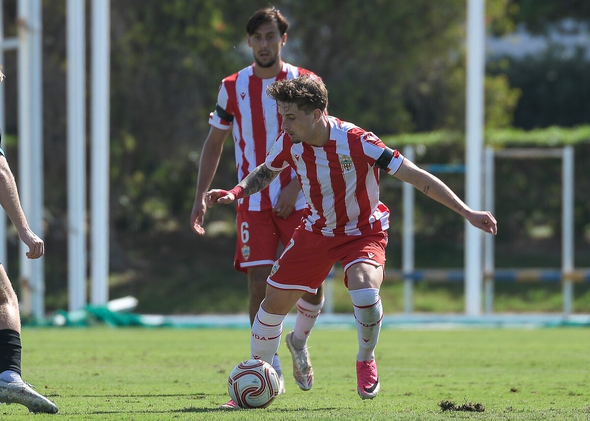 Dos futbolistas con camiseta rojiblanca a rayas en un campo de hierba; uno conduce el balón y otro le sigue detrás.