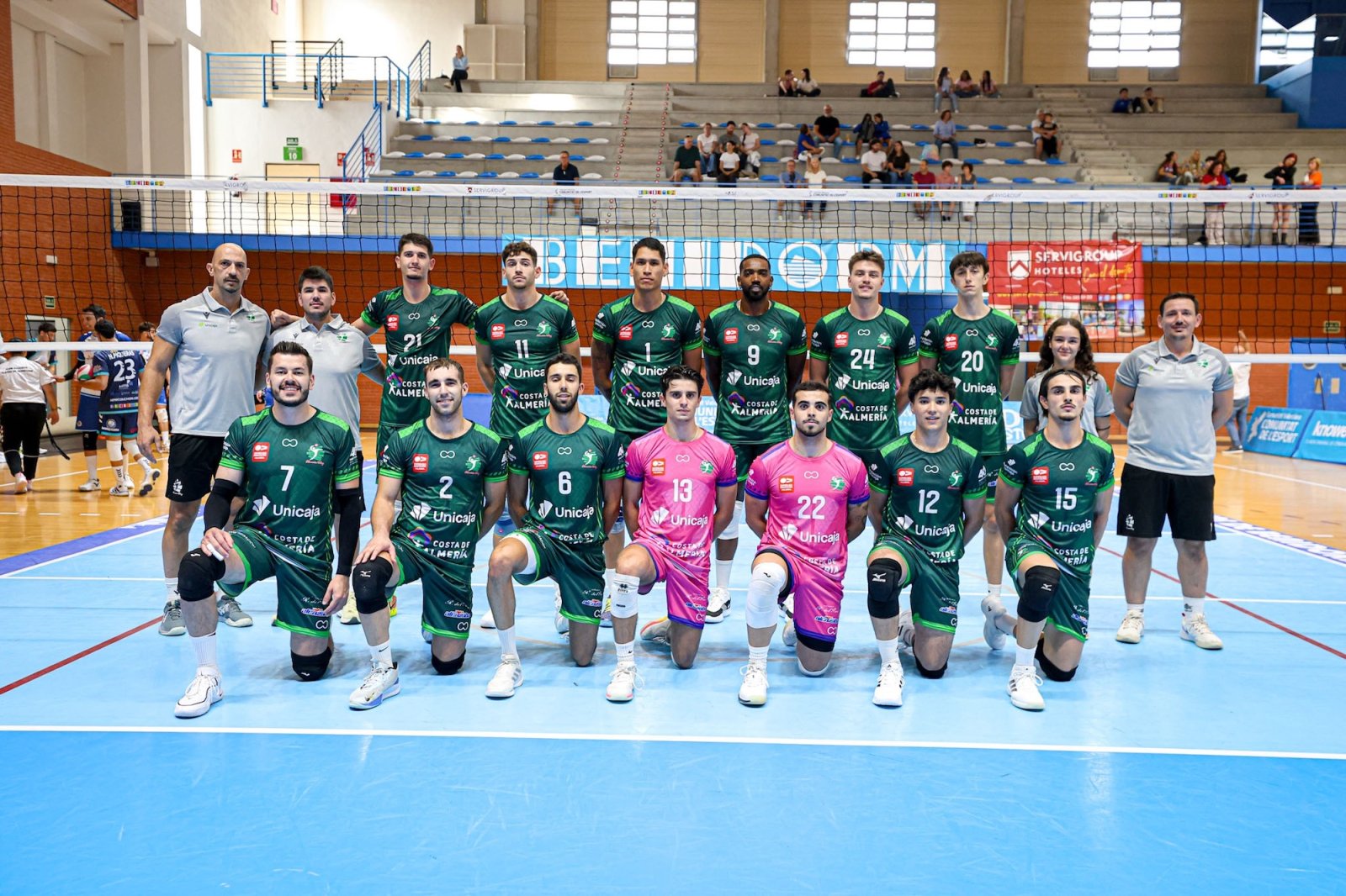 Equipo de voleibol posando en fila sobre pista azul junto a una red, con jugadores de verde y rosa y público en la grada.