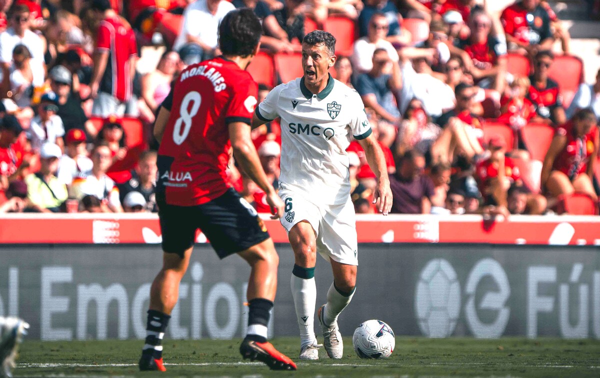 Un futbolista de blanco con el balón controlado ante un rival de rojo y negro, con público en la grada al fondo.