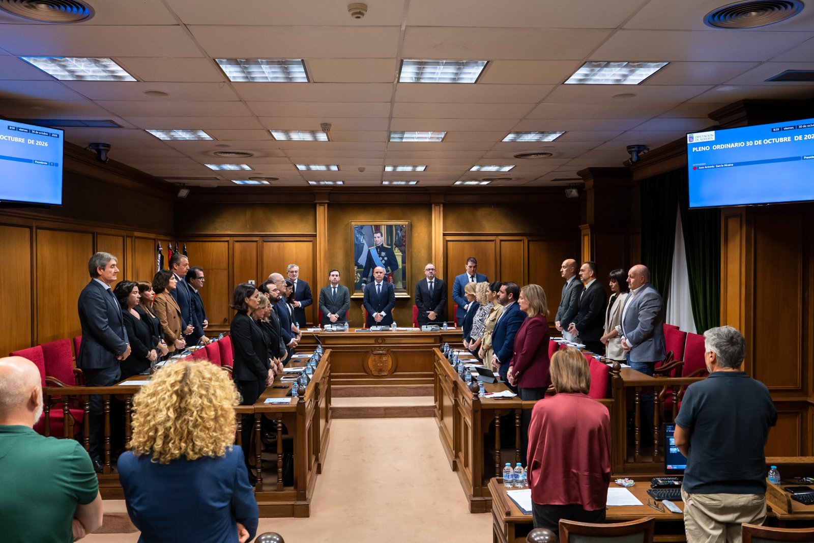 Personas de pie en un salón de plenos de madera con escaños rojos, pantallas azules y un retrato al fondo.