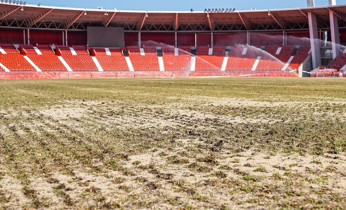 Césped en mal estado con calvas de arena en primer plano y gradas rojas vacías al fondo, con aspersores regando.
